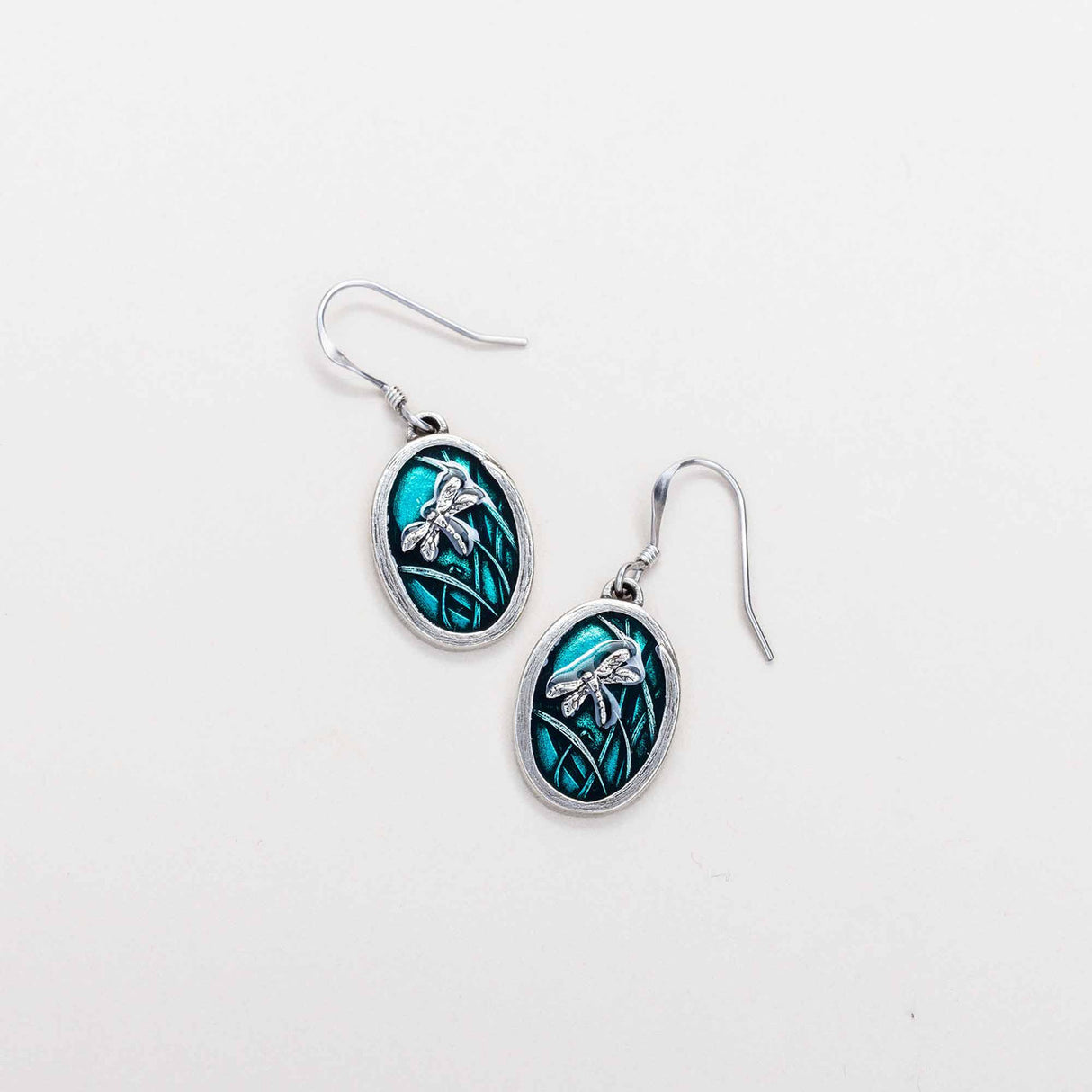 A pair of handcrafted oval shaped dragonfly earrings in teal displayed on a white background.