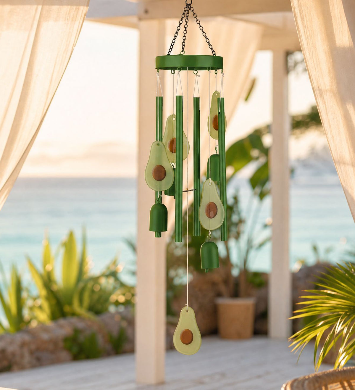 Green wind chime hanging outdoors under a white pergola with a beach and ocean in the background. The chime features avocado-shaped accents and green metal tubes.