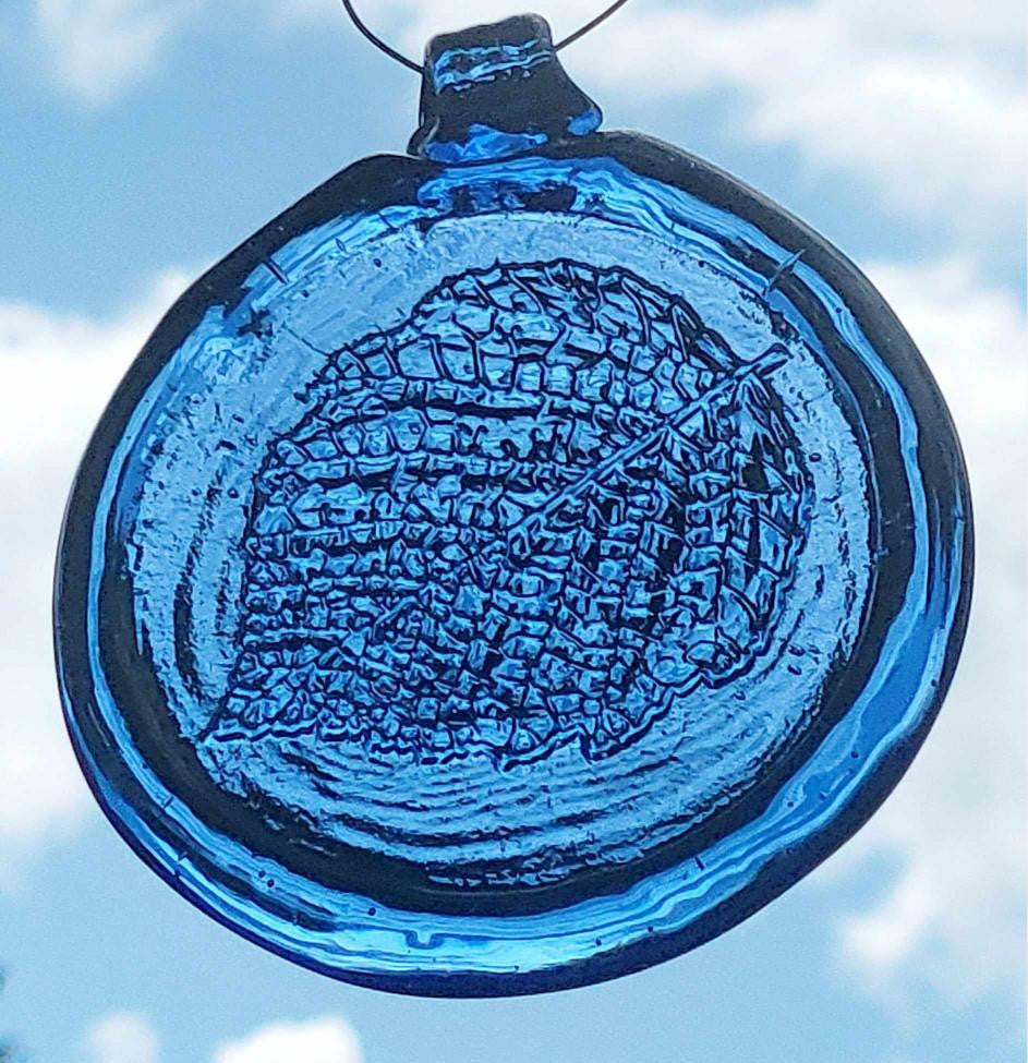 A blue circular suncatcher with a birch leaf pattern, hanging against a sky background.