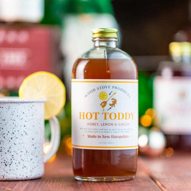 Hot toddy mixer bottle on a table top next to a steamy mug with a lemon wedge in it