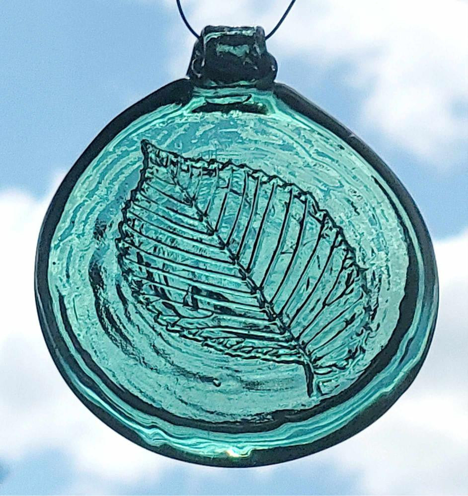 A hand-pressed glass suncatcher with an elm tree leaf pattern, in shades of blue and green, hanging against a sky background.