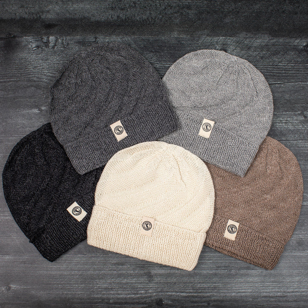 a grouping of neutral colored alpaca beanie style hats laid out on a gray wood floor - the hat colors include, from left to right, Charcoal, Gray, Cream, Silver, and Oatmeal