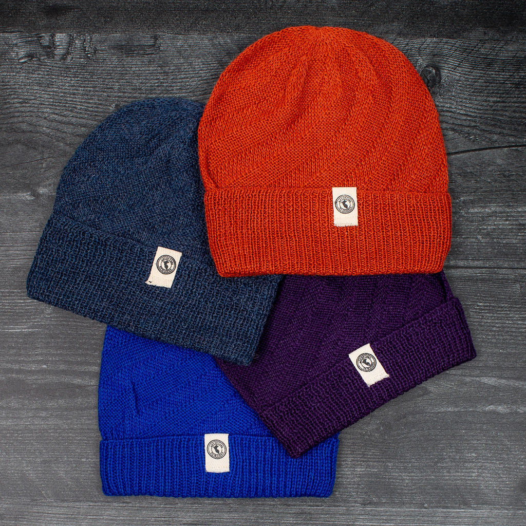 a grouping of colorful alpaca beanie style hats laid out on a gray wood floor. the hat colors include Denim, Rust, Blue, and Purple