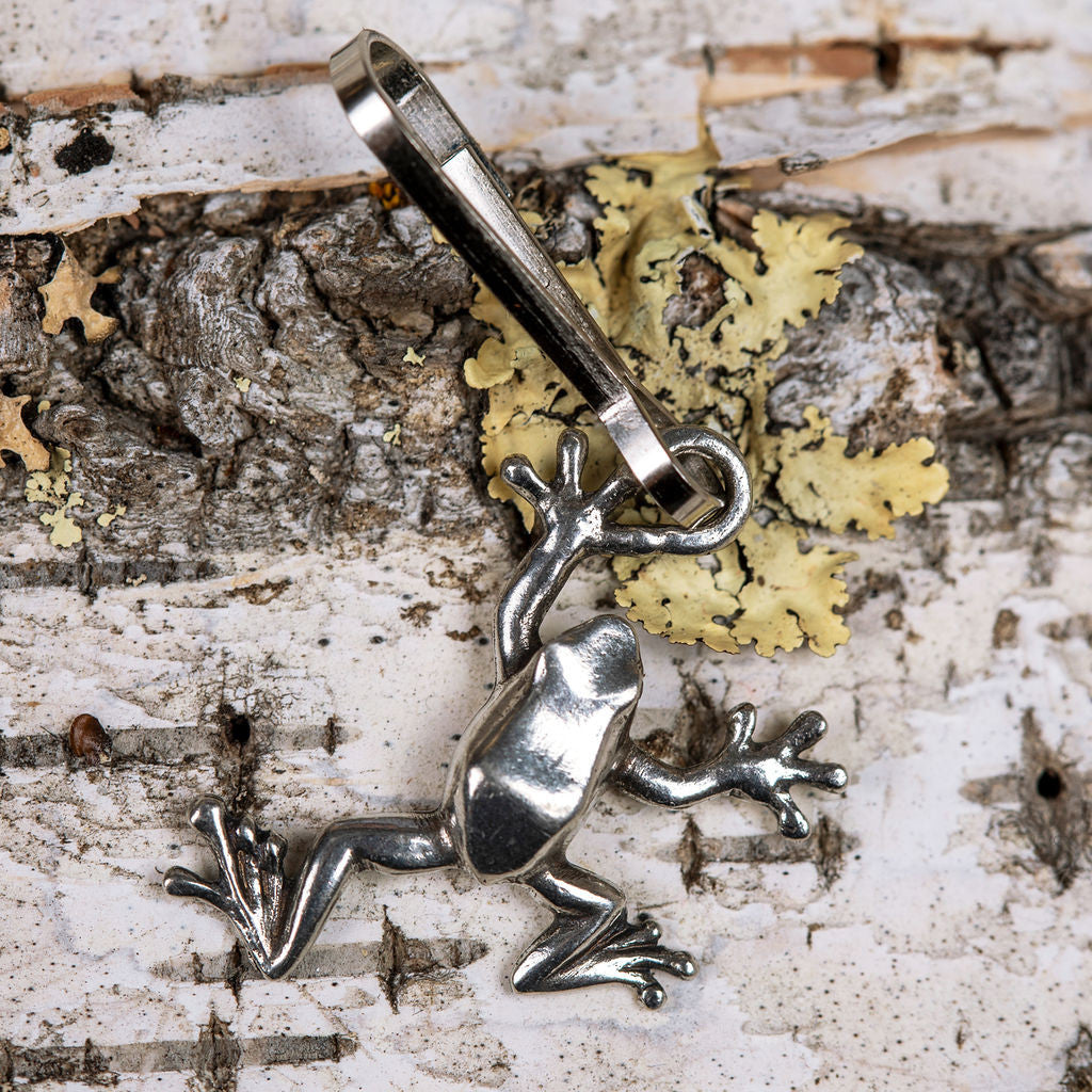 a handcrafted pewter tree frog zipper pull laid on a piece of white birch bark and posed as if it is climbing the tree