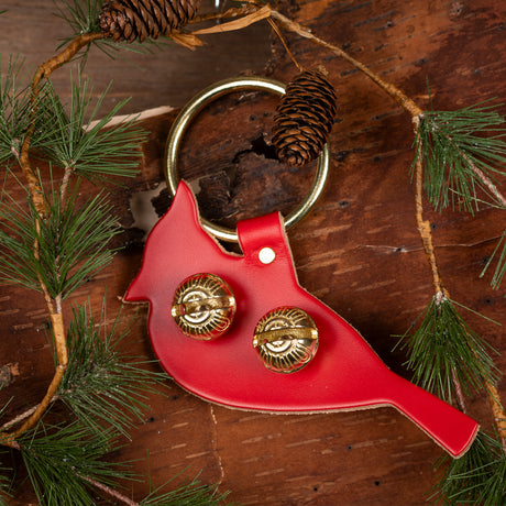 a door hanger bell in red leather stamped in the shape of a cardinal in profile laid on a natural bark background with evergreen accents