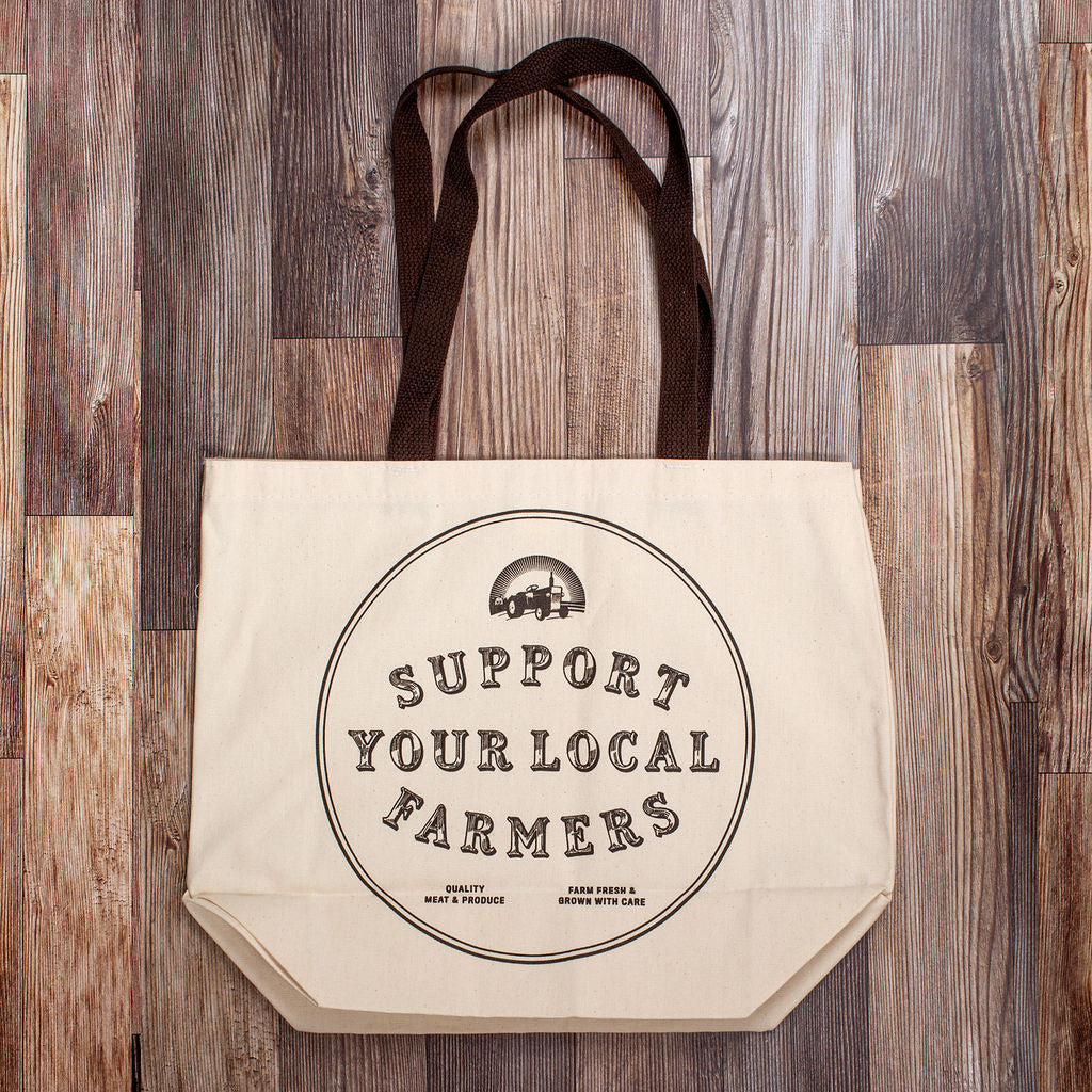 the back view of the canvas tote with brown strap against a dark wood grain background. within a circle the tote reads "support your local farmers" with a small tractor graphic above it