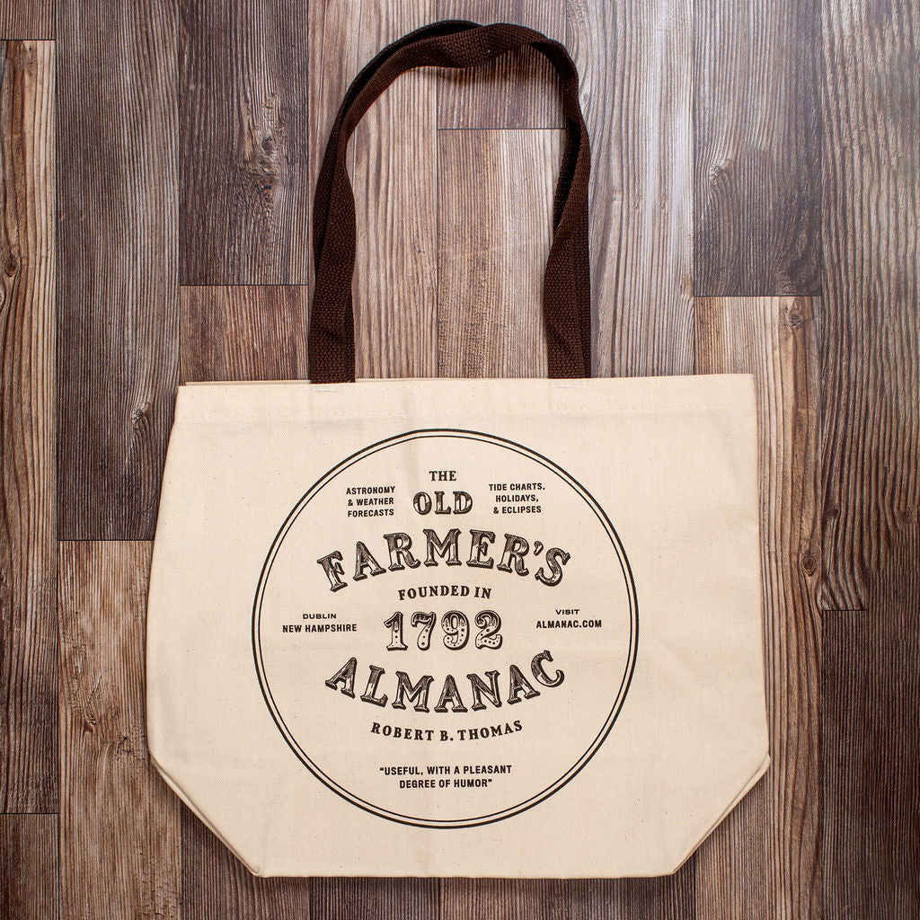 the front view of the canvas tote with brown strap against a dark wood grain background. The tote reads "Old Farmer's Almanac 1792" withing a circle graphic
