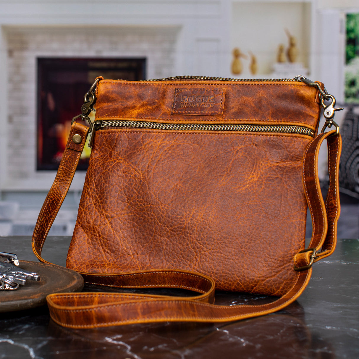 a nearly square, bison leather handbag sitting upright with the crossbody strap of the bag draped in front of it. The bag has a zipper across the front, upper portion of the bag and is a rich, copper colored leather with unique grain and subtle markings