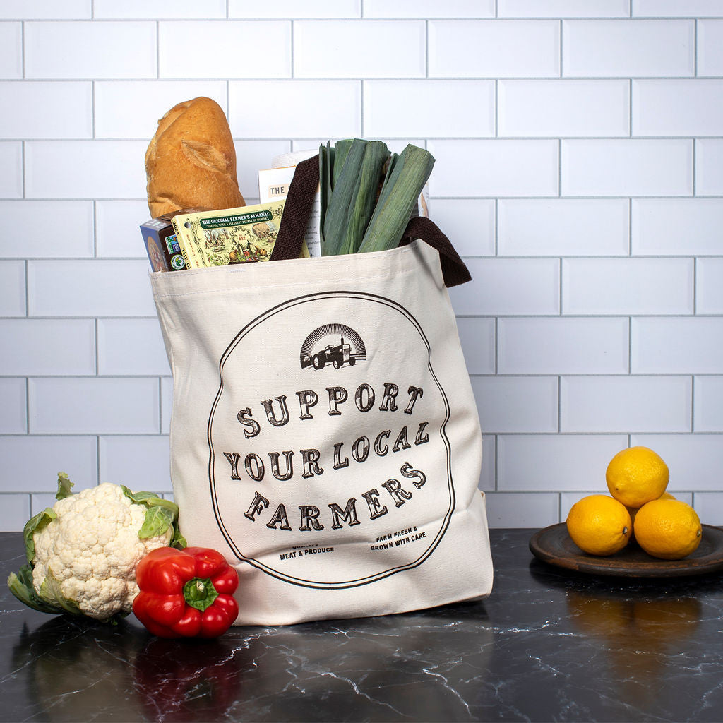The tote bag standing up on a dark marble countertop - the "Support your local farmers" side is facing front and the bag is filled with groceries and greens and next to lemons and other produce on the counter
