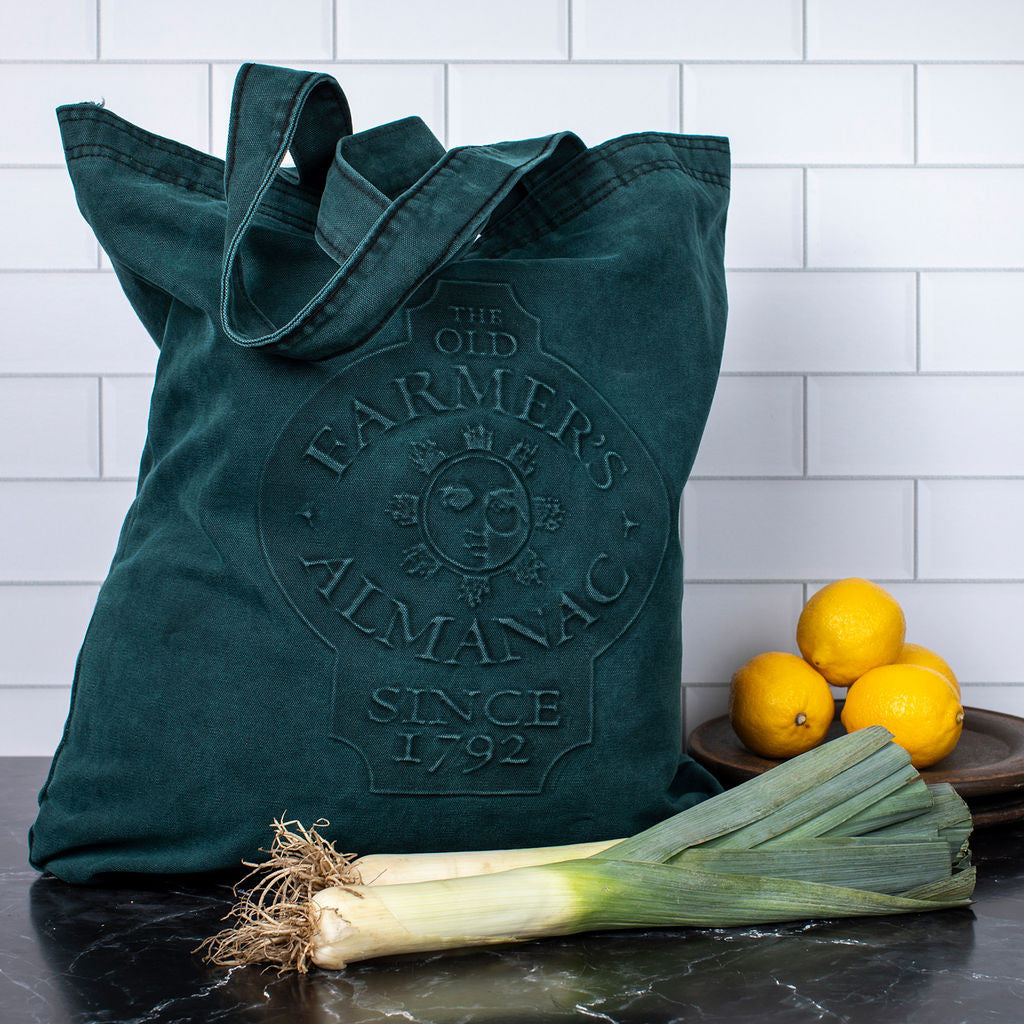 the Embossed Sun Face Tote filled and standing on a kitchen counter with leeks and lemons on the counter alongside it