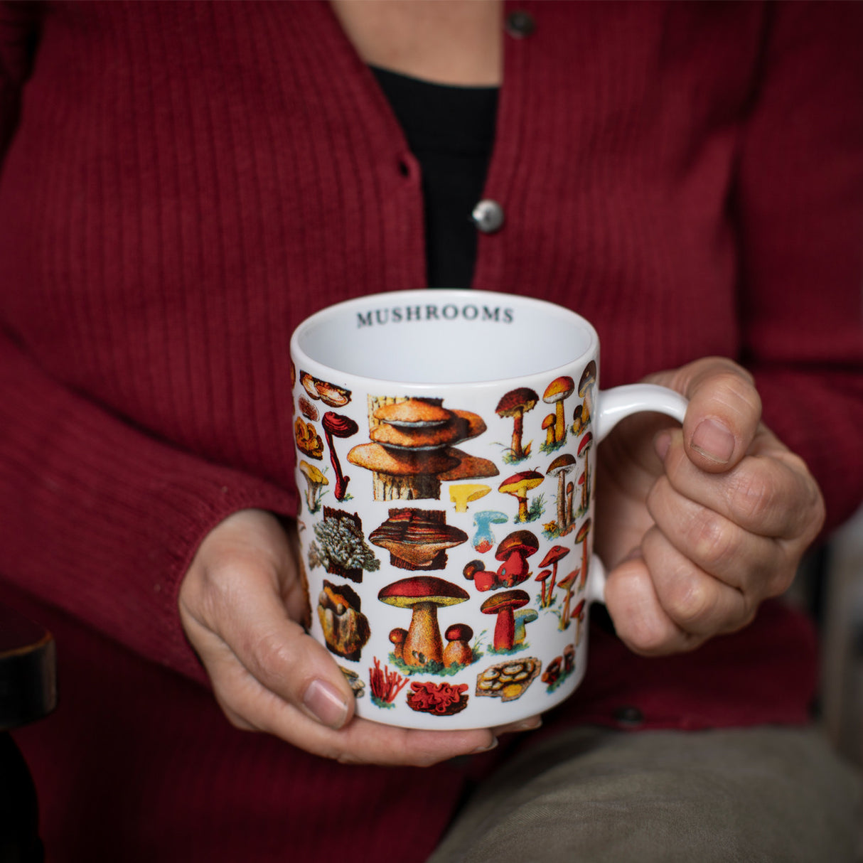 a seated model holds the mushrooms mug with the handle in her left hand. the inner lip of the mug revealing the text that reads "Mushrooms"