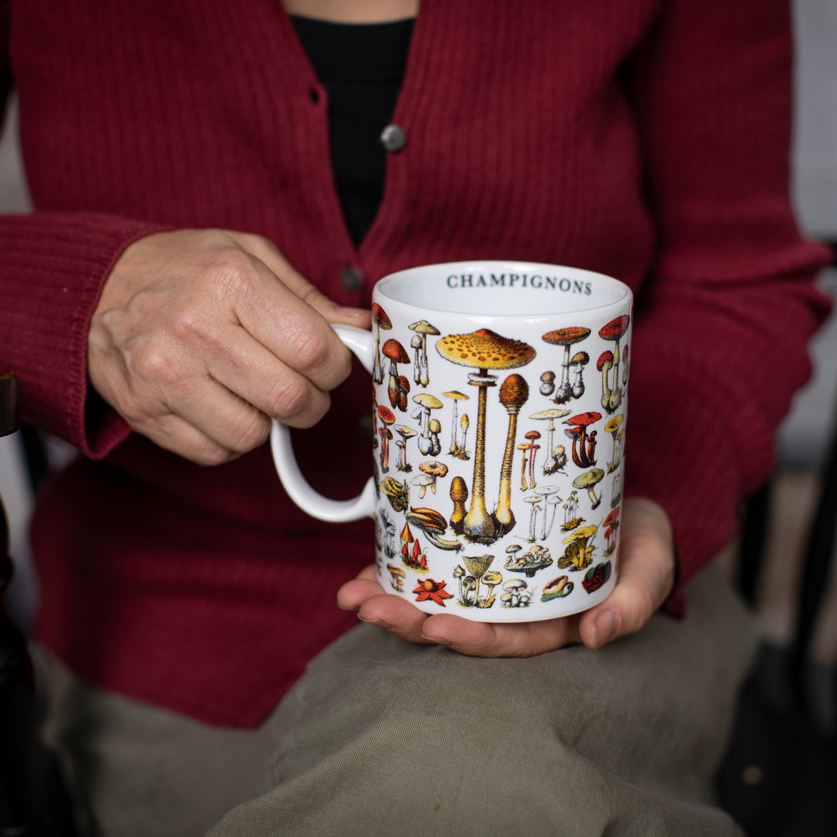 a seated model holds the mushrooms mug with the handle in her right hand. the inner lip of the mug revealing the text that reads "Champignons"