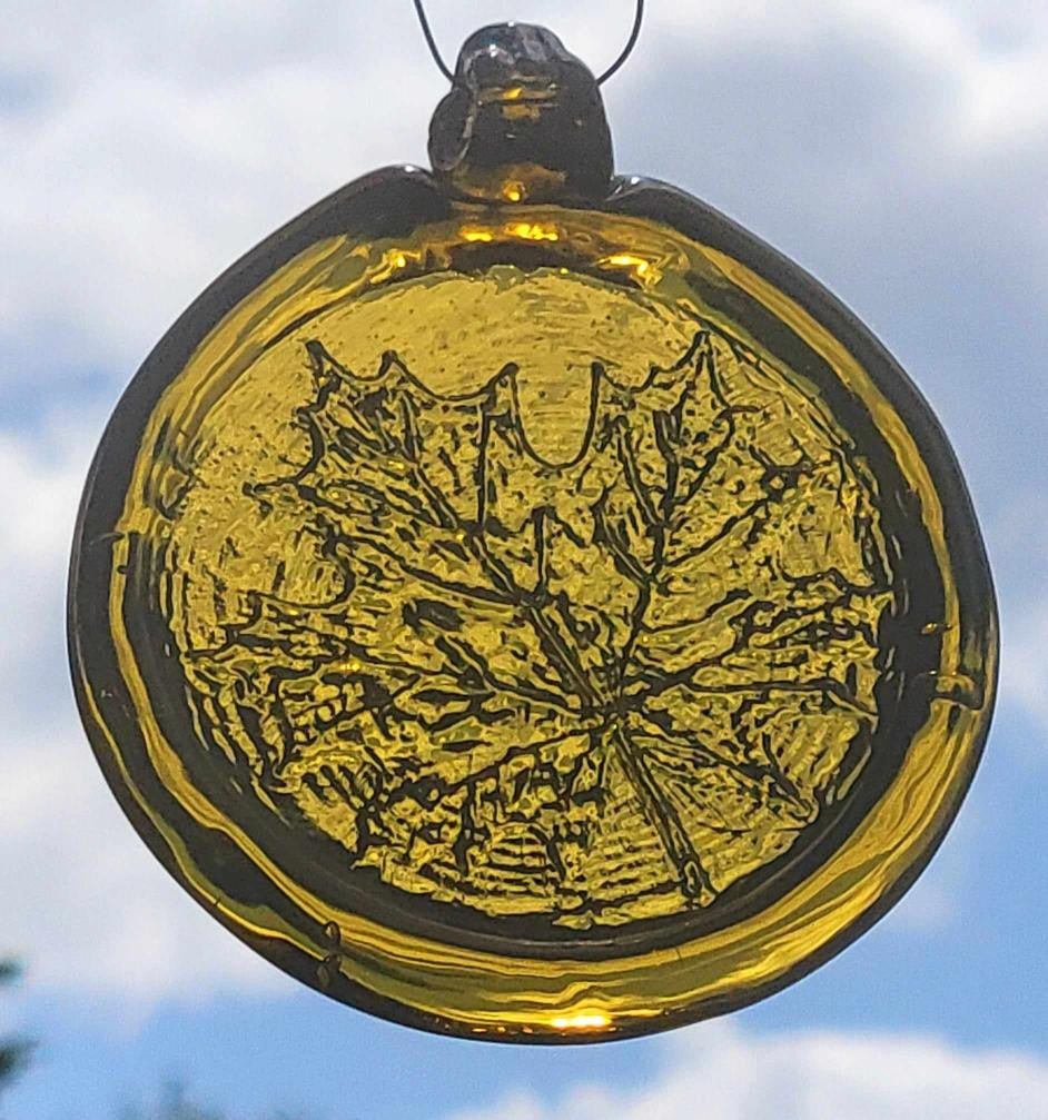 A hand pressed amber to yellow glass suncatcher with a maple tree leaf pattern, hanging against a sky background.