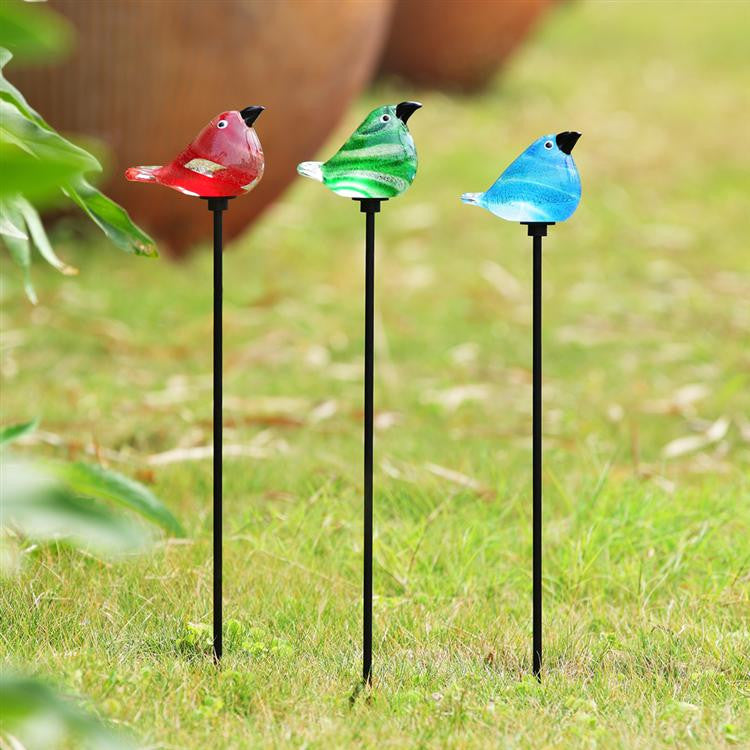 a lush lawn featuring the 3 glass bird stakes in a row: red, green, and blue shown from left to right