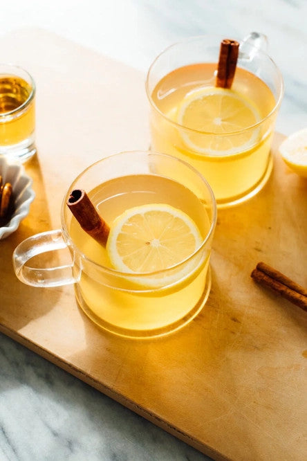 a tabletop with several glasses of warm amber liquid in clear rocks glasses and garnished with lemon slices and cinnamon stick