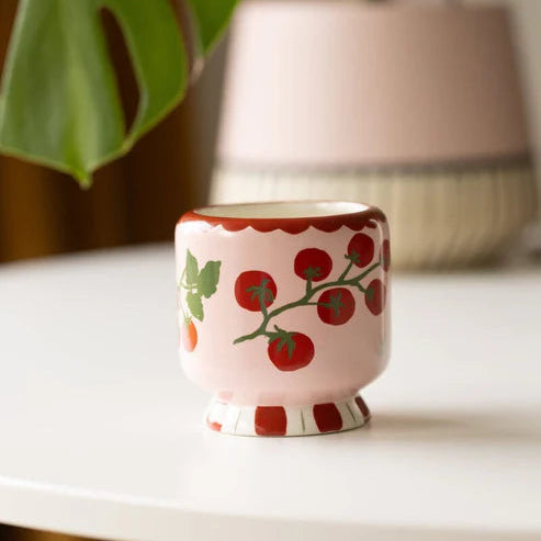 Small ceramic vessel with tomato vine design on a white surface, with a plant and lamp in the background.
