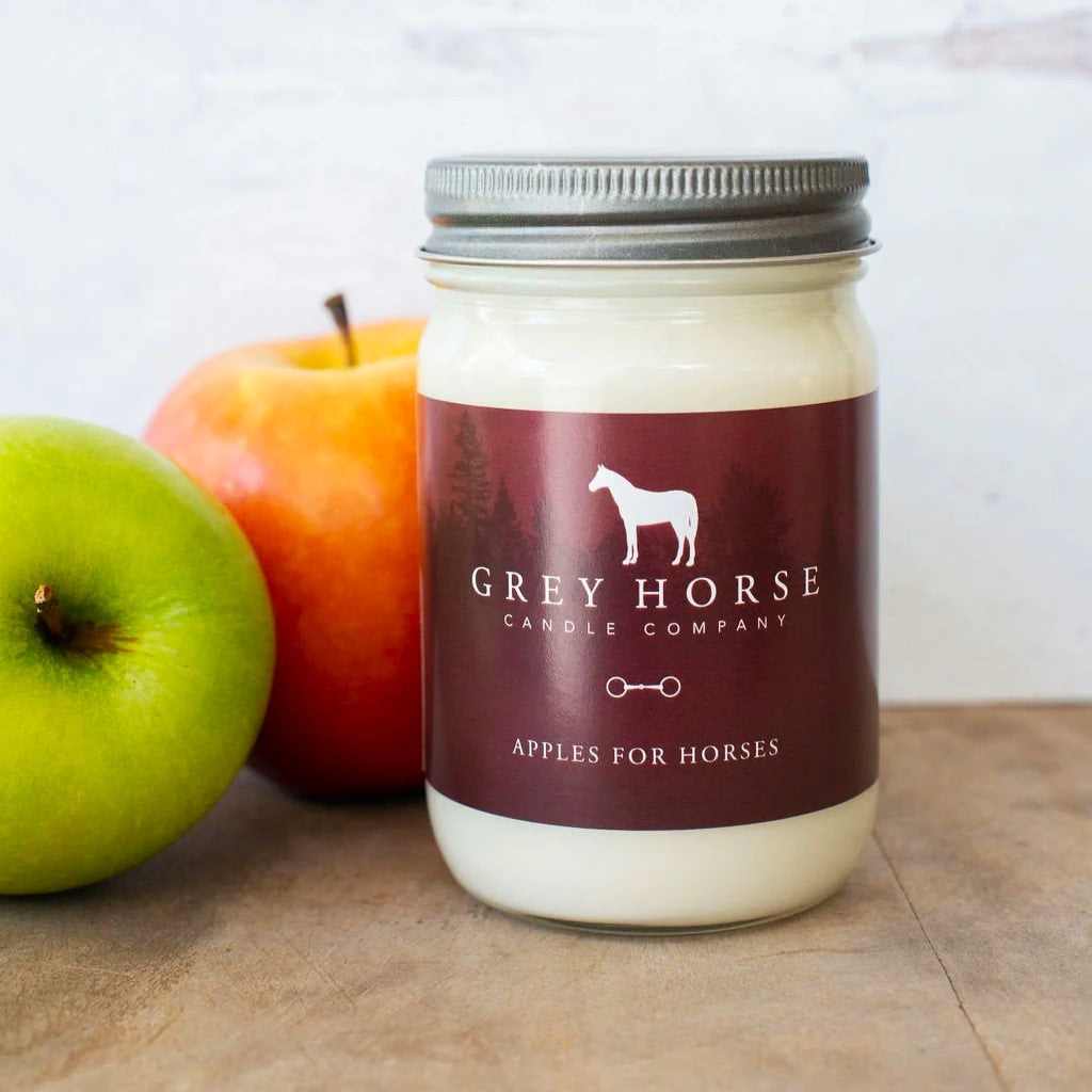 a hand poured candle in a clear glass canning jar with a deep red label that reads "Grey Horse Candle Company, Apples for Horses" sits on a wood surface alongside a red apple and a green apple