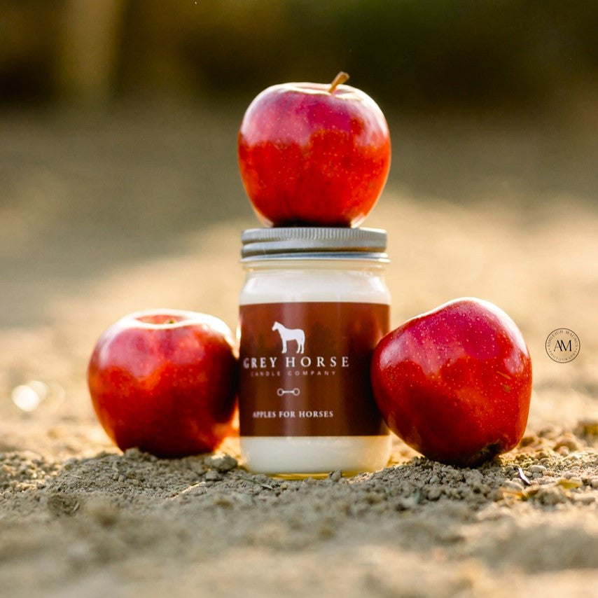 a glass candle jar on a dirt surface surrounded by red apples