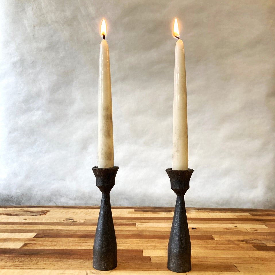 a pair of forged candle holders with lit white taper candles on a light wood surface