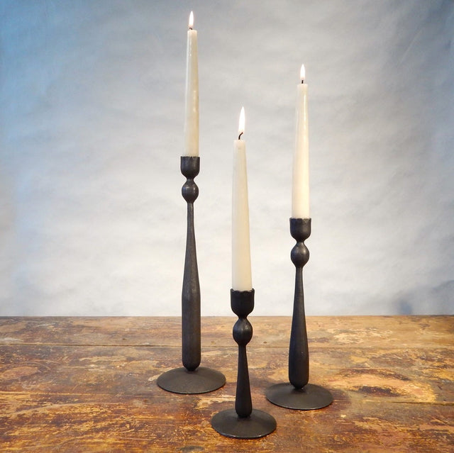 3 iron candlesticks of varied heights holding lit off-white taper candles sitting on a wood surface