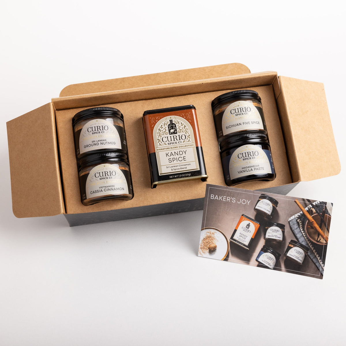 A curated gift box of Curio Spices containing four spice jars and a box of Kandy Spice, displayed in an open brown cardboard box with a 'Baker's Joy' card on a white background.