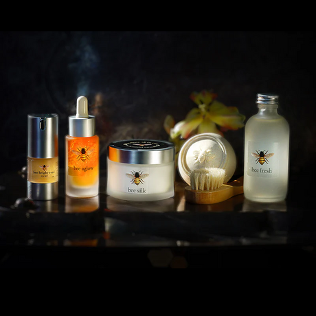 The Bee Renewed I skin care regimen line up of products, from left to right, Bee Bright-Eyed, Bee Aglow, Bee Silk, Bee Soothed, and Bee Fresh against a black background