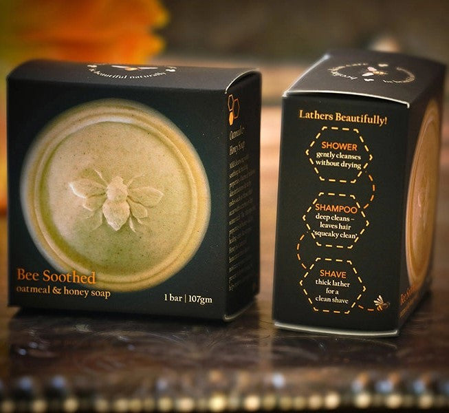 the Bee Soothed soap bar in its packaging showing the honey bee imprint on the bar of soap and suggested uses on the side of the box: Shower, Shampoo, and Shave