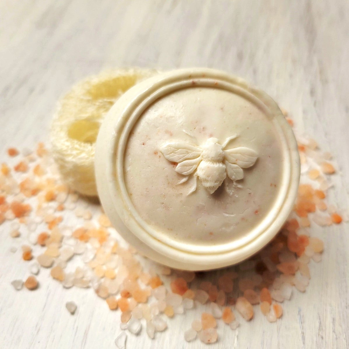 a close up of the Bee Heeled scrub bar which shows the detailed bee imprint on the circular bar of soap, the exfoliating loofa imbedded in the soap, and a sprinkling of pink himalayan sea salt around the soap