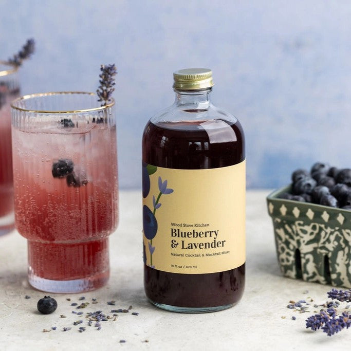 a tabletop scene with a bottle of Blueberry & Lavender mixer in the center and surrounded by glasses with brilliant purple beverages and a container of fresh blueberries
