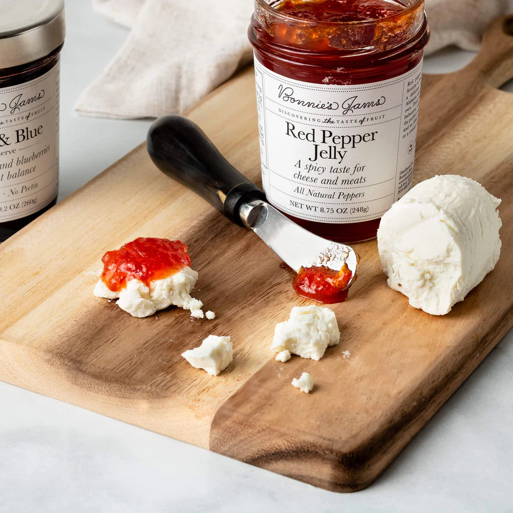 Bonnie's Jam Red Pepper Jelly on a wooden board with cheese and a spreader.