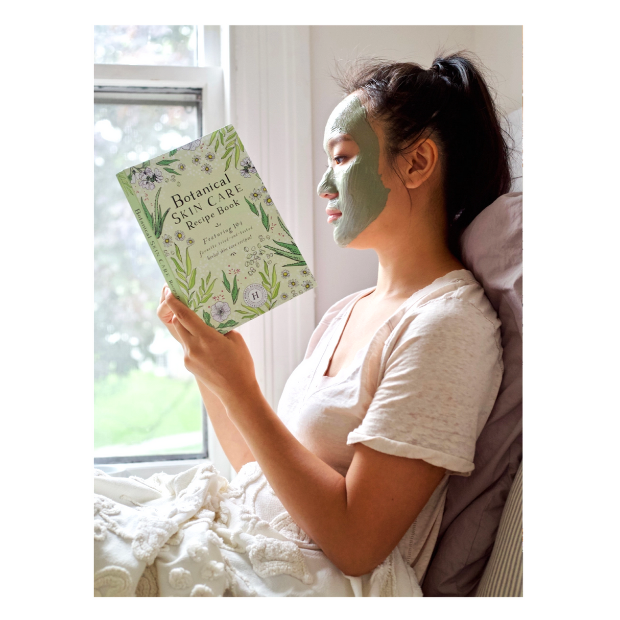 a model wearing a green face mask sitting and reading the Botanical Skin Care Recipe Book