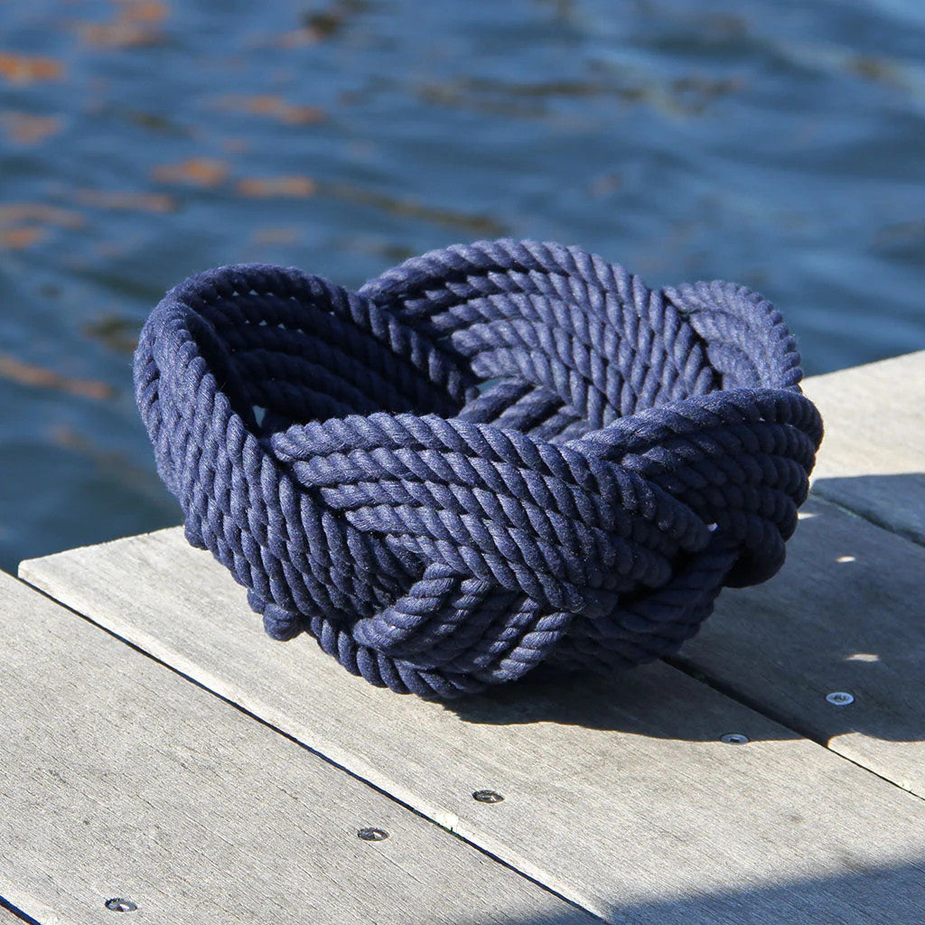 a hand woven cotton rope bowl in navy blue resting on a dock with a view of the water