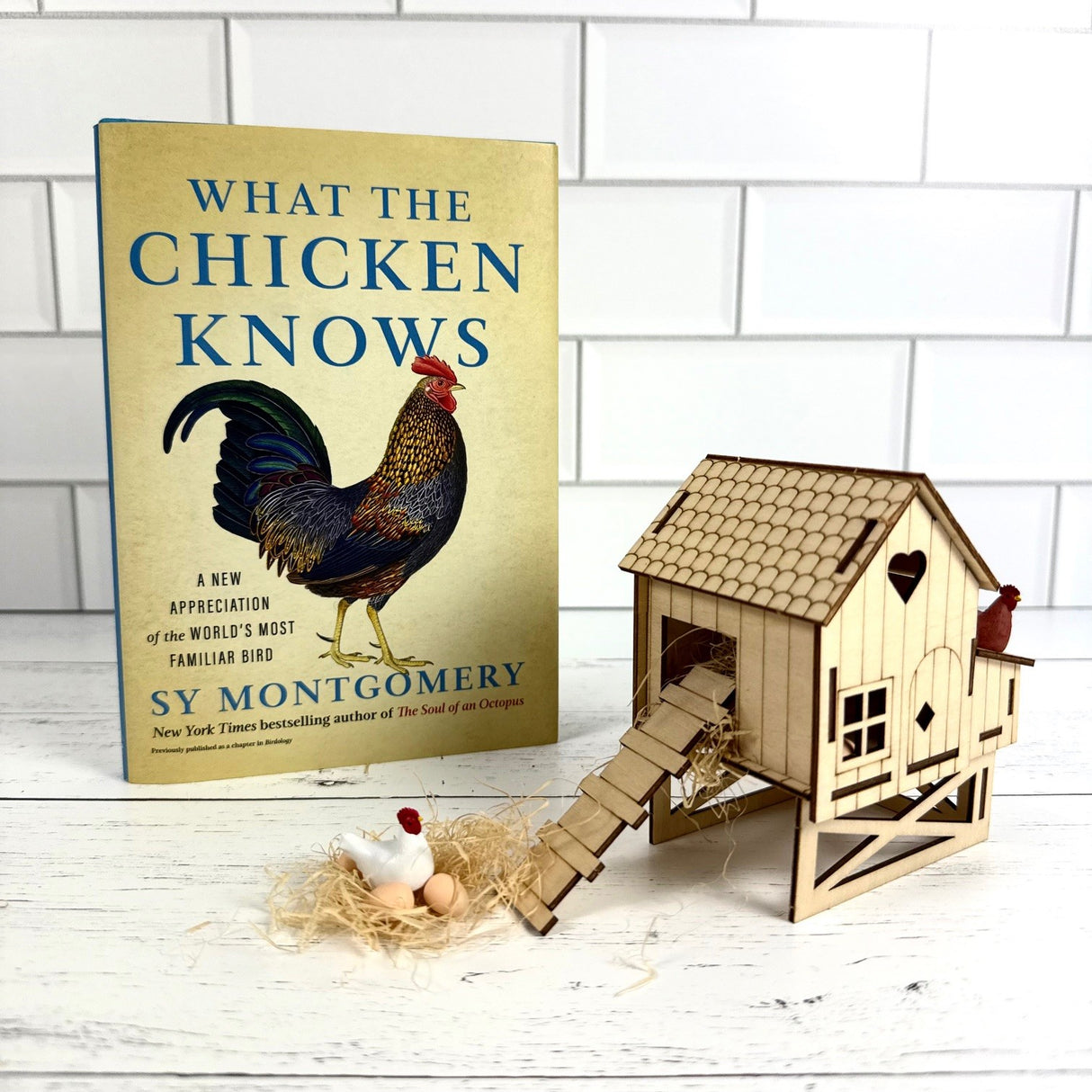 Book titled 'What the Chicken Knows' by Sy Montgomery with a wooden chicken coop model on a white surface.