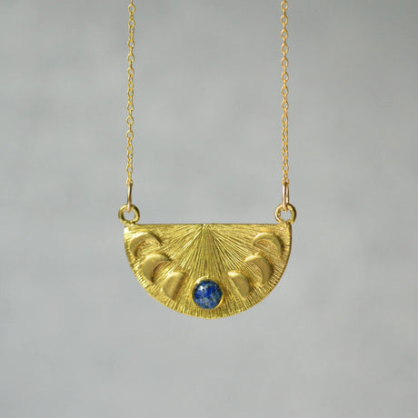 a brass cast pendant features a delicate half moon shape, with each phase of the moon represented in intricate detail. At the center, the full moon is highlighted by crushed lapis lazuli. The pendant hangs from a gold chain