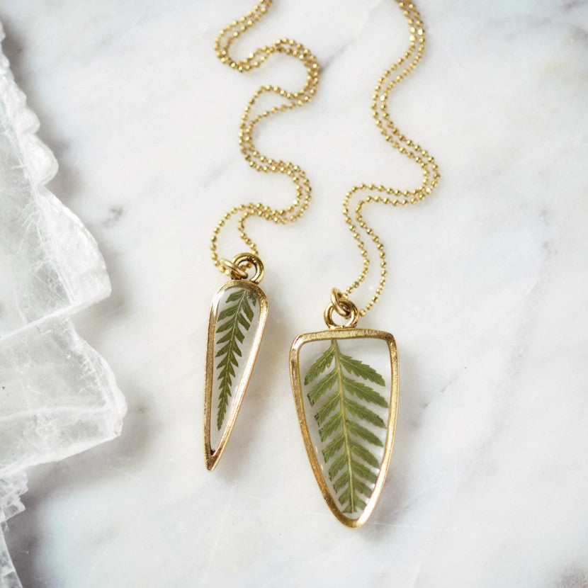 a necklace featuring a preserved fern pressed inside clear resin, framed by a delicate 24kt gold-plated arrow-shaped pendant with a gold bead chain laid on a white marble surface