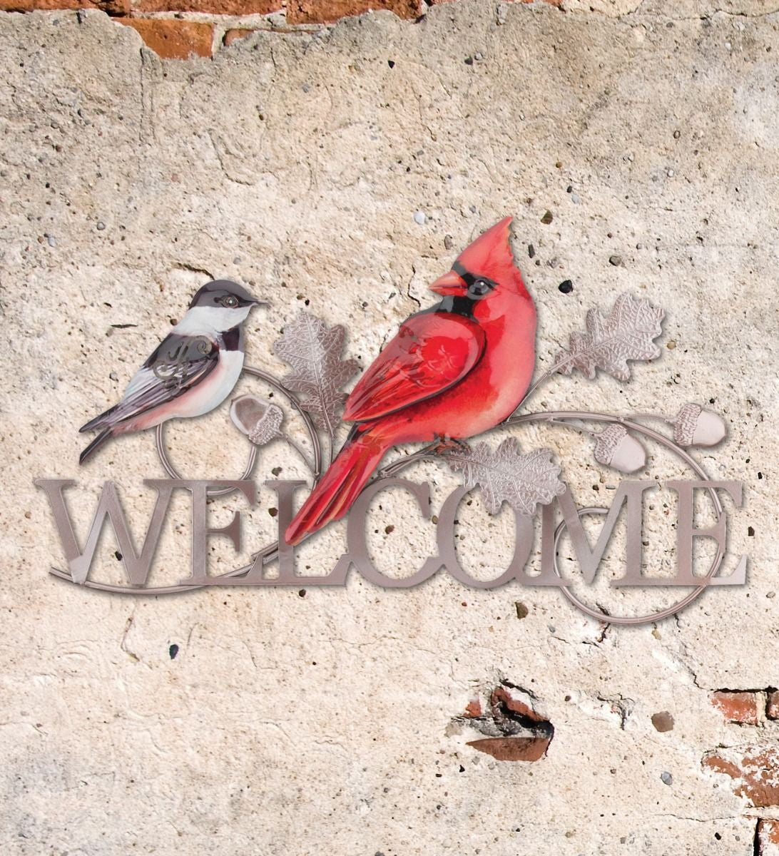 a silver welcome sign in silver metal with a realistic cardinal and chickadee perched atop the sign installed on a beige stucco wall