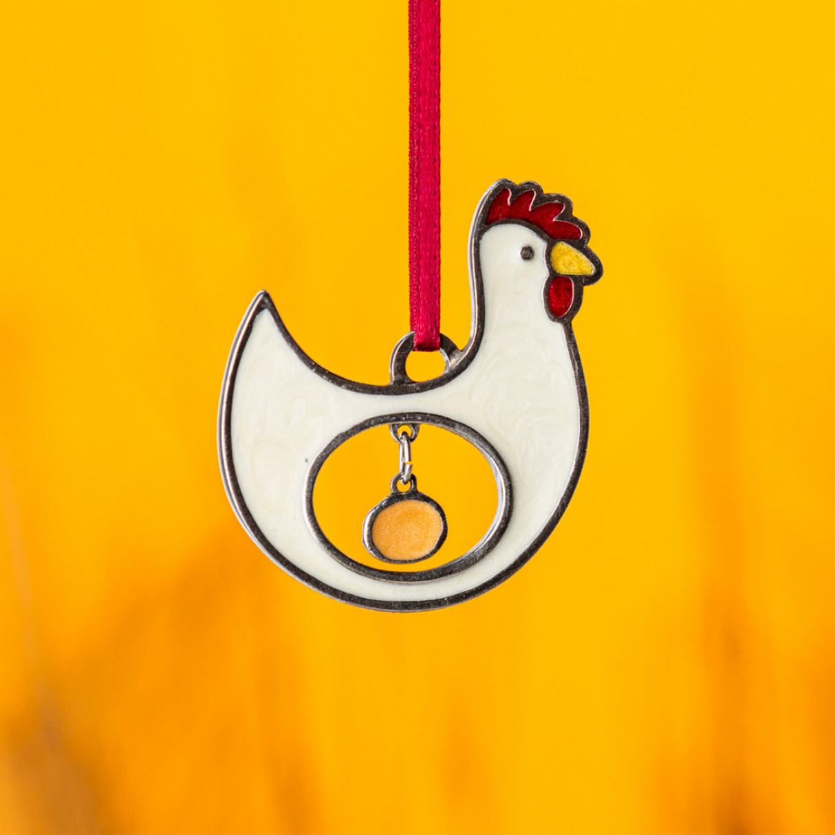 pewter ornament of a chicken with a hollow center where an egg is hanging within the center against a yolk-yellow background