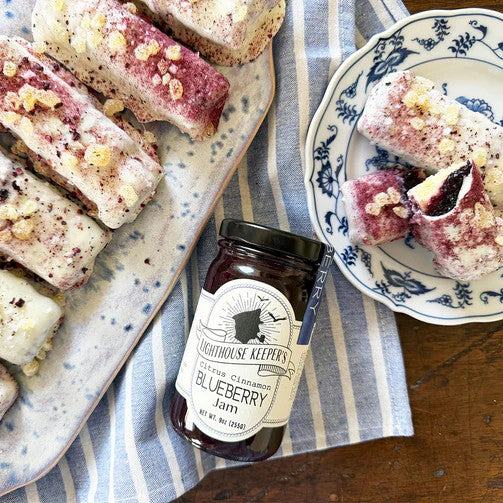 a jar of Blueberry jam laying on a table alongside a platter of sliced pastry and a serving of pastry on a plate on a classic white and blue striped cloth