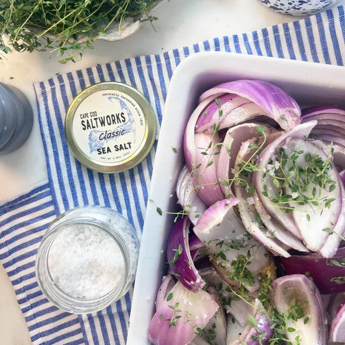 an open jar of sea salt alongside a baking dish with vibrant red onions and herbs ready for roasting