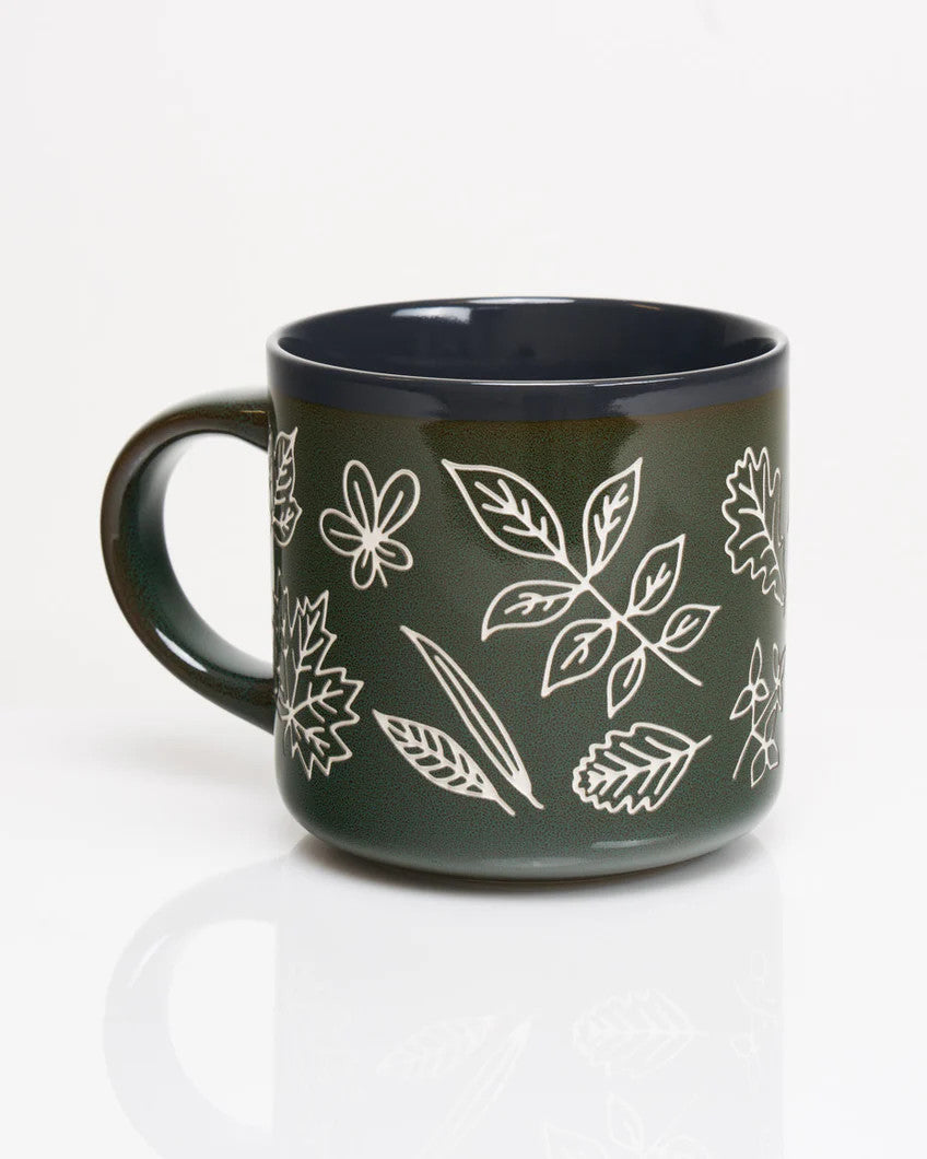 a forest green coffee mug with various leaf prints in white showing the outlines and details of various leaves in white with a vintage feel