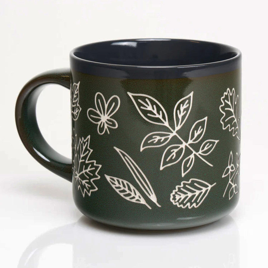 a forest green coffee mug with various leaf prints in white showing the outlines and details of various leaves in white with a vintage feel