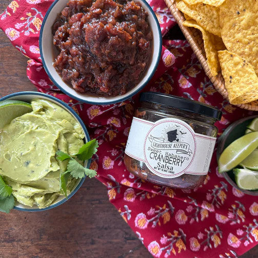 a tabletop with a salsa spread with a jar of cranberry salsa, a dish filled with cranberry salsa, a bowl of tortilla chips, and a dish of guacamole