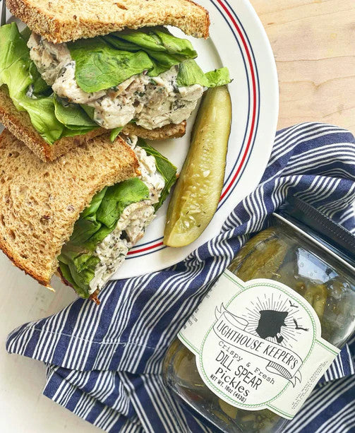a tabletop spread with a chicken salad sandwhich on a plate with a pickle spear next to a jar of lighthouse keeper's pantry dill spear pickles