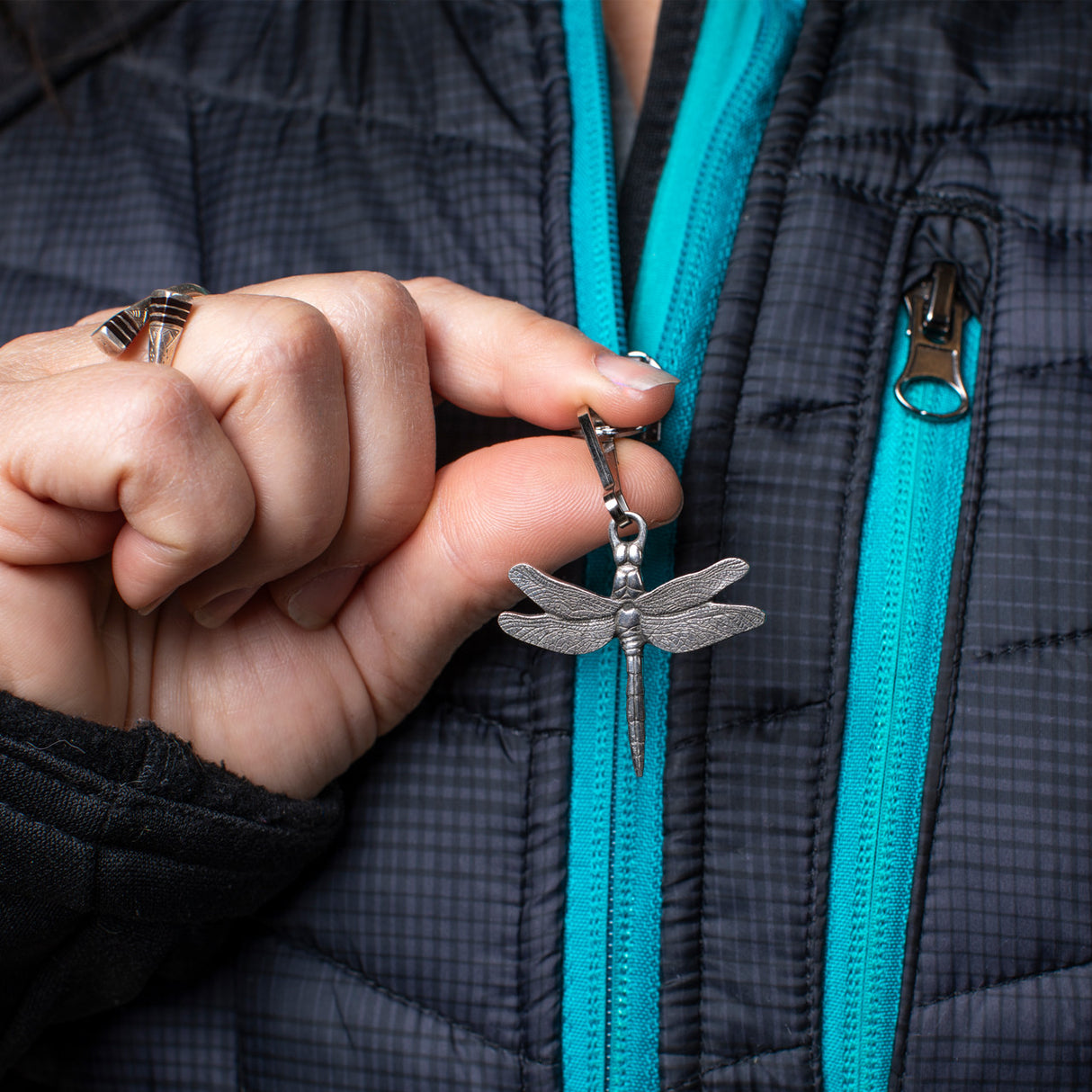 close up of a model holding her dragonfly zipper pull between her thumb and forefinger - the zipper pull is attached to the zipper on her black coat with turquoise accents