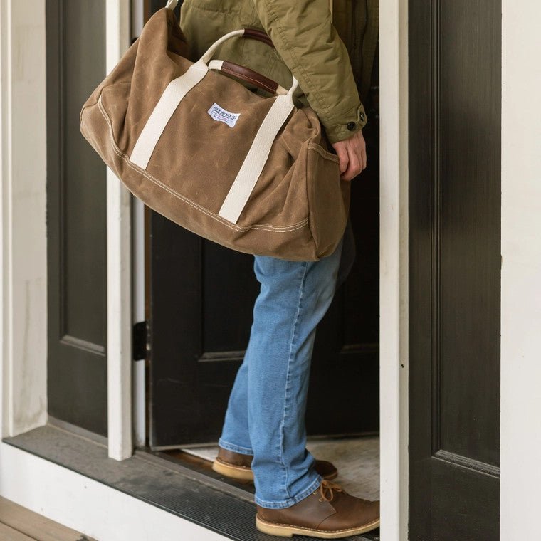 a model standing in a doorway with their brown waxed canvas duffle bag slung over one shoulder