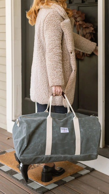 a model opening a door gripping her gray waxed canvas duffle bag by the leather handles