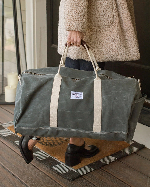 a close up of a model gripping the gray waxed canvas duffle bag by the leather handles