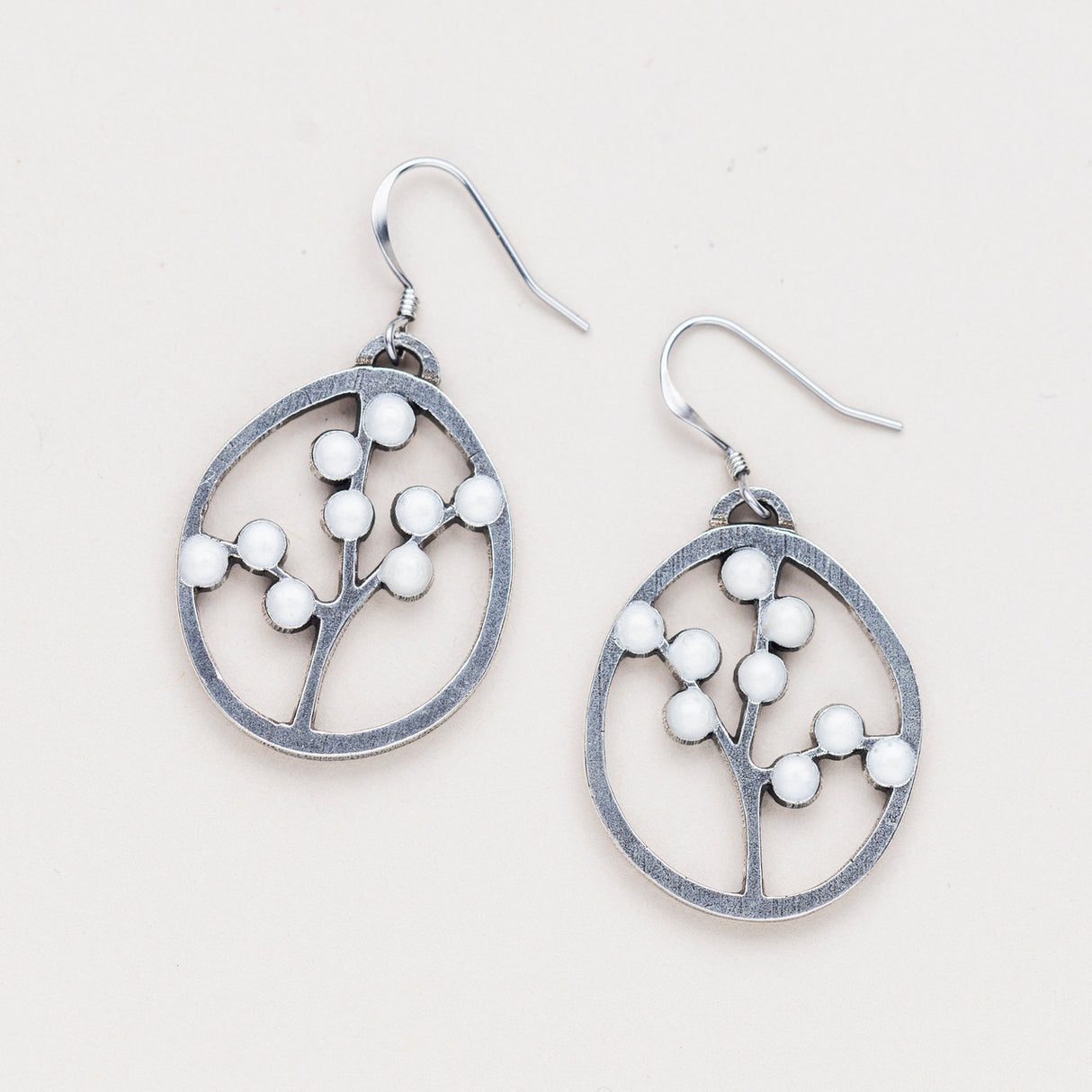 a pair of oval pewter earrings with branches inside adorned with white beads to represent blossoms