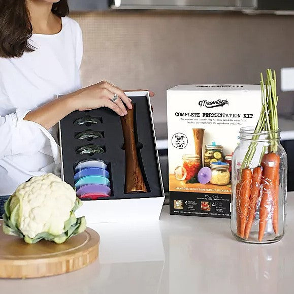 a model at a countertop holding the opened package of the mason jar fermentation tool kit with raw cabbage on the counter along with a bunch of carrots in a mason jar