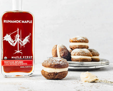 a bottle of festivus infused maple syrup on a countertop next to some gingerbread whoopie pies, highlighting the syrup's potential for use as a baking/flavoring ingredient